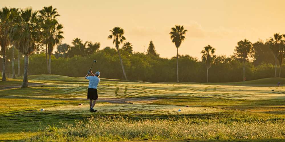 A golfer swinging a club at sunrise, surrounded by lush greens and palm trees at South Padre Island Golf Club.